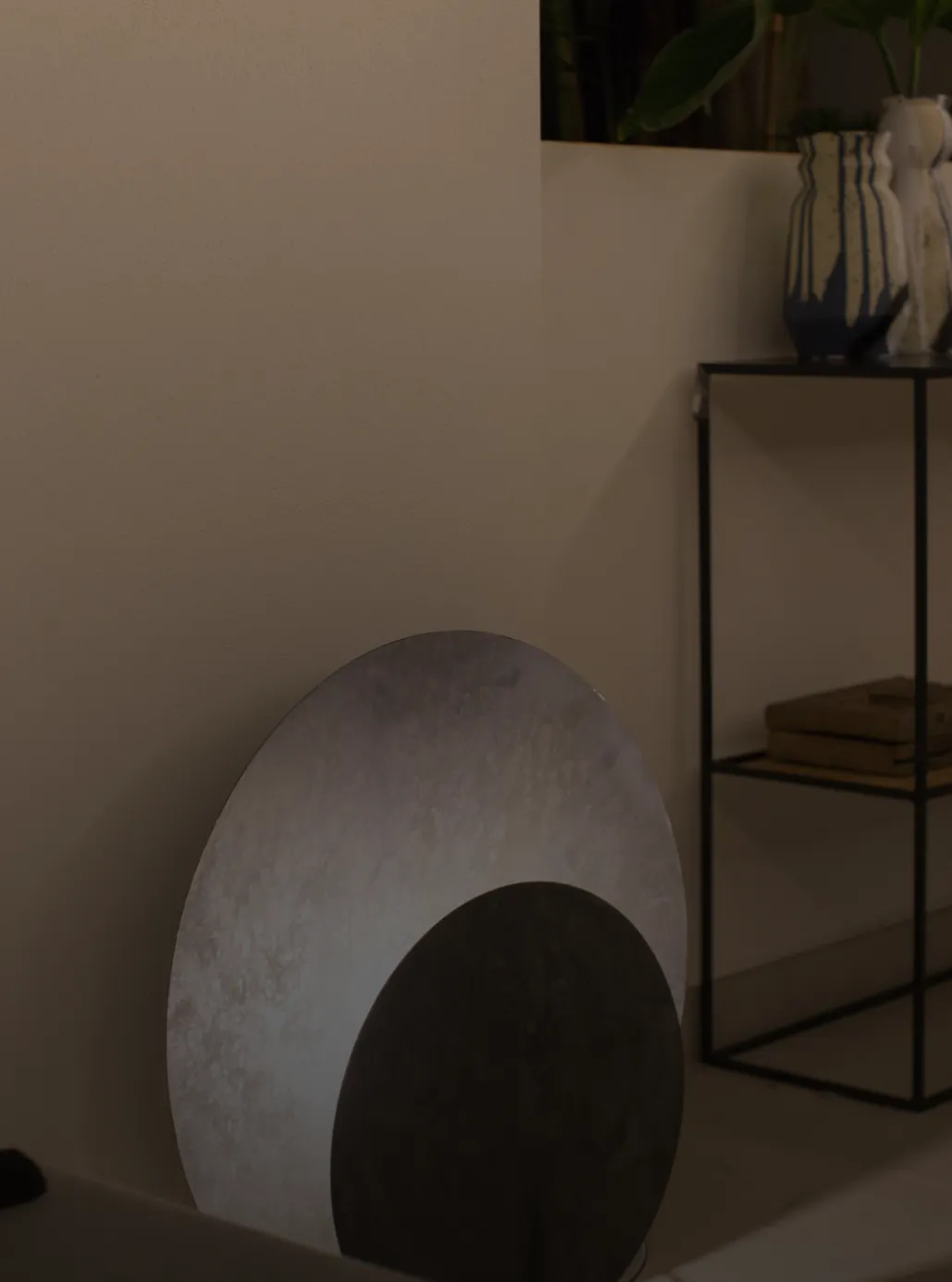Round alabaster mirror leaning against a wall, beside a console with ceramic vases