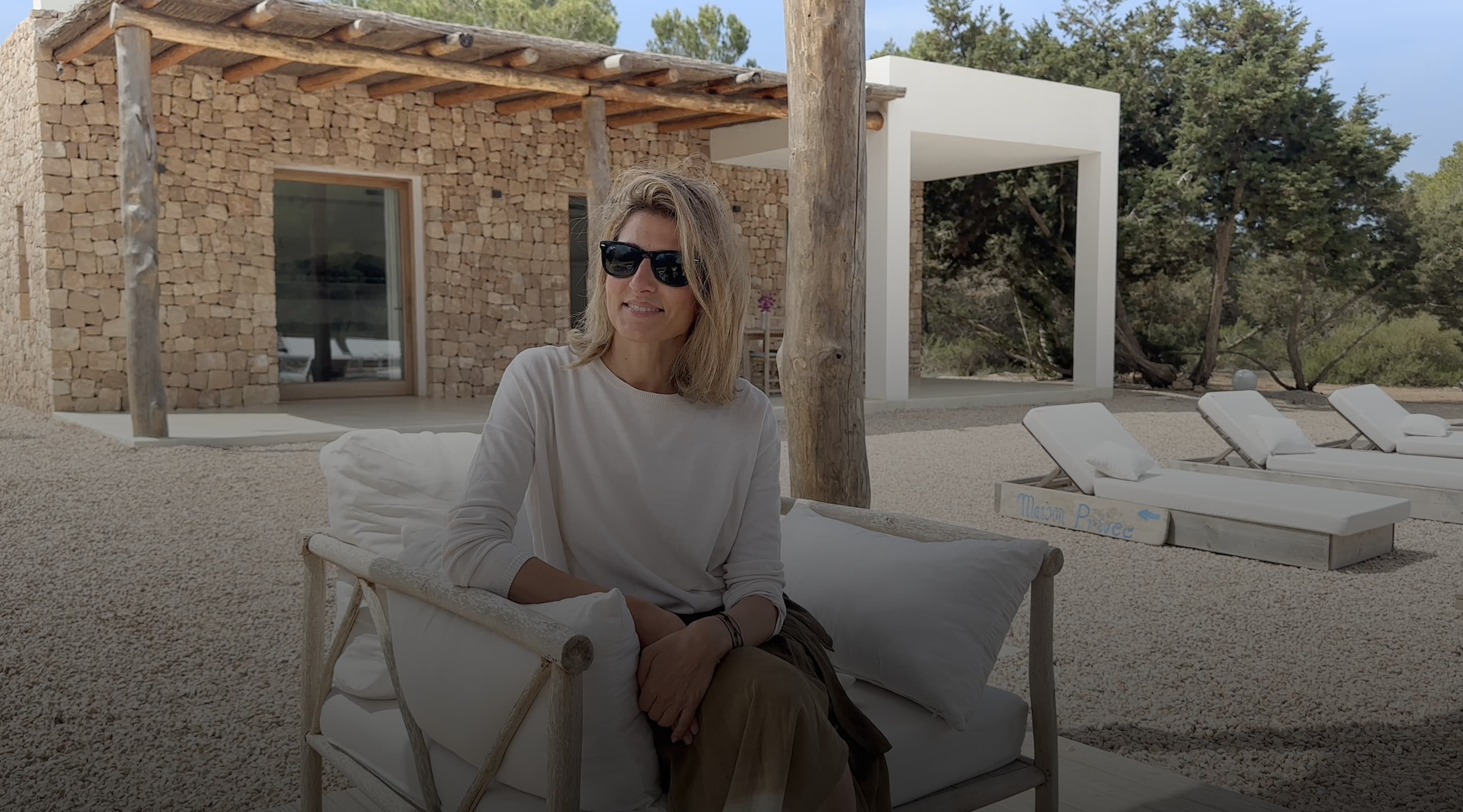 A woman sitting in an outdoor armchair in front of a stone house with a pergola