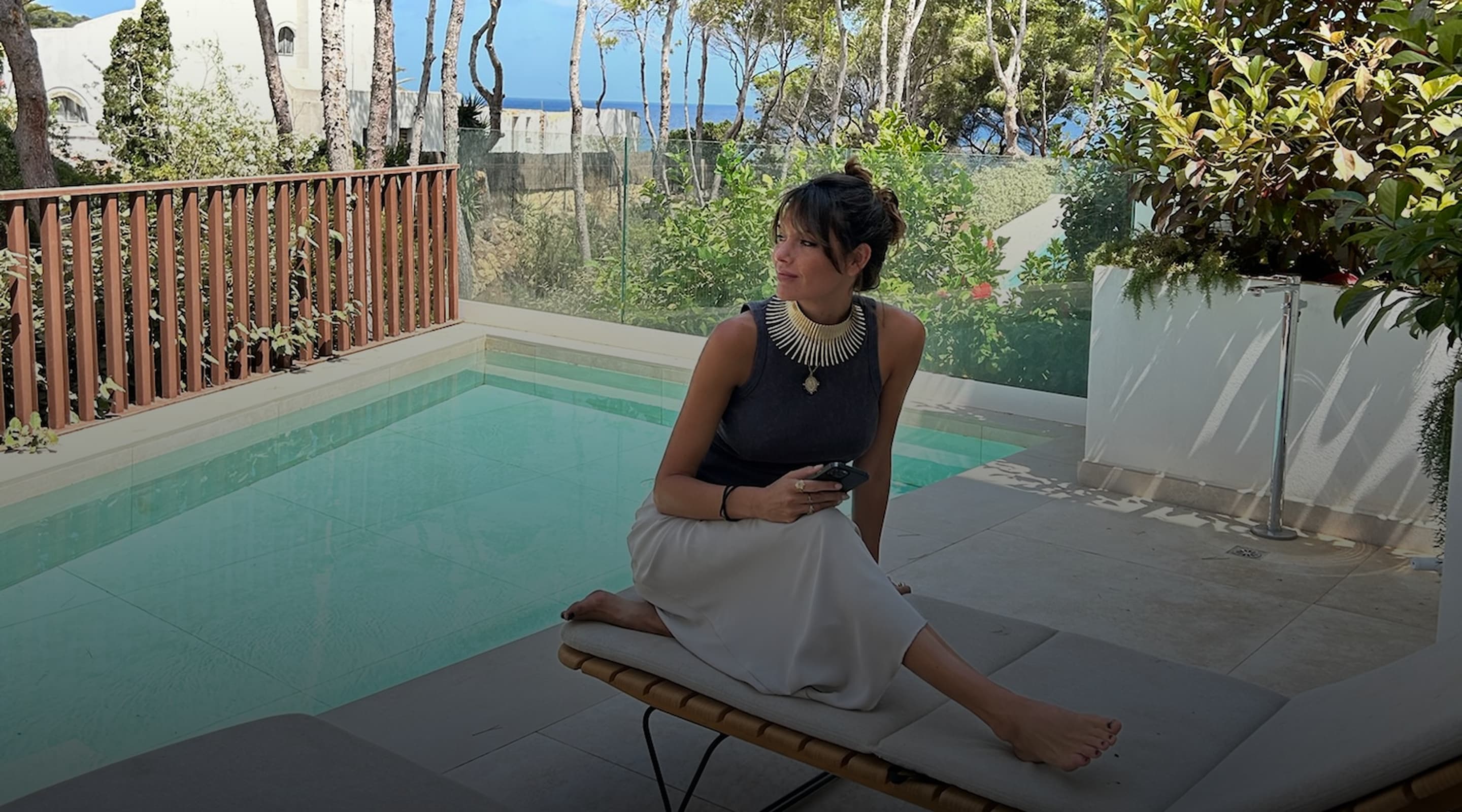 A woman sitting beside a small pool, with pines and the sea behind